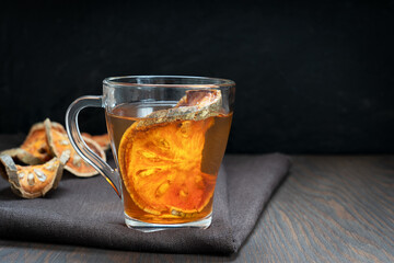 Traditional Thai healing matoom tea made of dried bael fruit slices, also known as wood apple, in transparent glass cup on brown towel on dark wooden background. Image with copy space, horizontal 