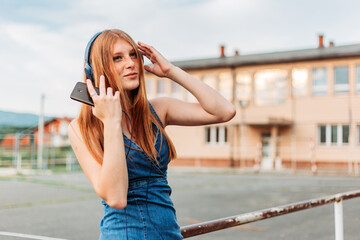 Ginger teen streaming music on her phone. Headphones on her head, school yard