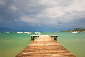 Fototapeta premium A footbridge leads to the troubled Lake Garda in Italy. The blue-green water surface is churned, lots of waves. Boats dance in the water. The sky is blue, a storm is coming. Long exposure time.