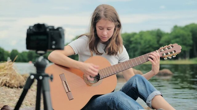 young woman blogger sitting near lake with a guitar in his hands in front of the camera