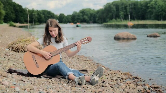 Young girl of European appearance playing an acoustic guitar on the shore of the lake, hobby concept, musical creativity