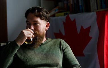 Young stylish handsome bearded Canadian man enjoying whiskey with a Canadian flag behind him