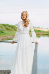 Fototapeta na wymiar Blonde hair bride in white silk long lace dress standing on the terrace with lake view