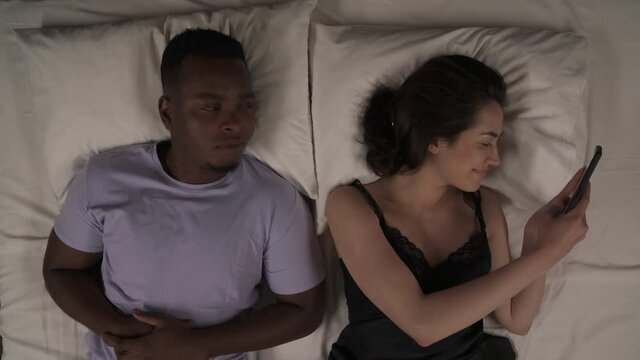 Top view of mixed-race couple in bed at night. White girl staring at her mobile phone and smiling. Black man peeking at screen, making facepalm gesture