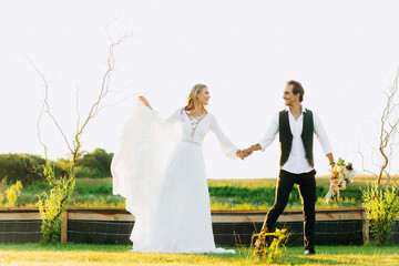 Stylish couple of happy newlyweds walking in the field on their wedding day with bouquet