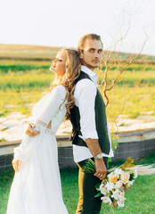 Stylish couple of happy newlyweds walking in the field on their wedding day with bouquet