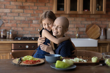 Loving cute little girl child hug sick cancer patient bald mother cooking healthy food salad in kitchen together, caring small daughter embrace show love support to ill mom, feel grateful thankful