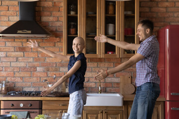 Happy young Caucasian couple with sick cancer patient woman wife have fun dance together enjoy weekend in kitchen at home, smiling man feel playful optimistic with ill bald hairless female spouse