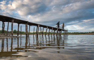 Naklejka premium Happy couple man and woman with bicycles on the pier in the lake against the backdrop of the cloudy sky.
