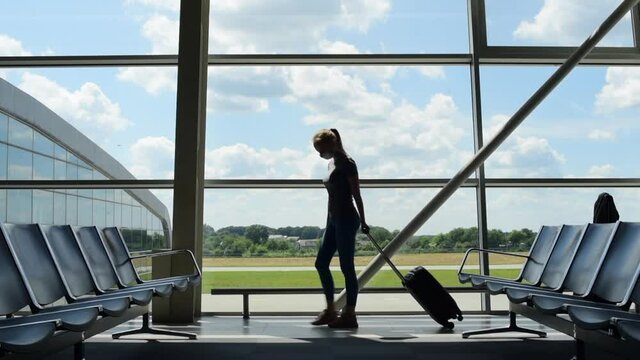 Woman tourist wearing medical protection face mask walks with luggage in empty airport terminal during COVID-19.