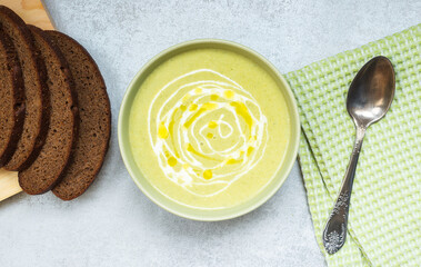 Flavorful, homemade broccoli cream soup, with olive oil and cream in a green Cup, on a light gray table.There is bread, a napkin, and a spoon next to it. Top view, close-up