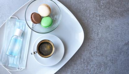 A Cup of coffee, macaroon cakes, a medical mask and antiseptic on a white platter on a table. The concept of opening a cafe after the end of the coronavirus pandemic, a new normal concept. Copyspace