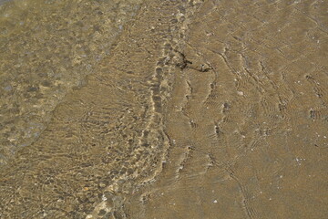 Mehrere kleine Wellen am Meeresufer werden von der Sonne beleuchtet und bilden eine Textur aus feine Schatten auf dem hellen feinen Sand mit wenige kleine Muscheln und Meeresalgen.