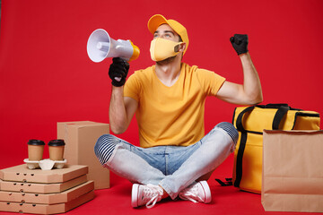 Delivery employee man in yellow cap face mask gloves tshirt sit at thermal takeaway food bag backpack pizza boxes work courier service during quarantine coronavirus covid-19 isolated on red background