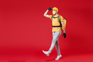 Fototapeta na wymiar Full length delivery employee man in yellow cap face mask gloves t-shirt uniform thermal food bag backpack work courier service during quarantine coronavirus covid-19 virus isolated on red background.