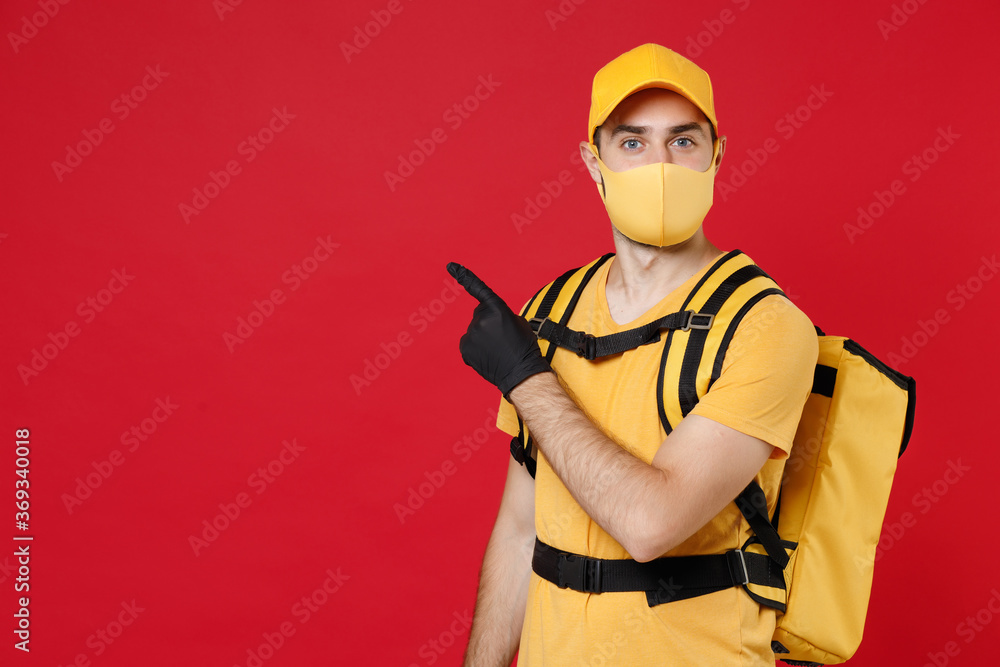 Wall mural Delivery employee man in yellow cap face mask gloves t-shirt uniform thermal bag backpack with food work courier service during quarantine coronavirus covid-19 virus isolated on red background studio.