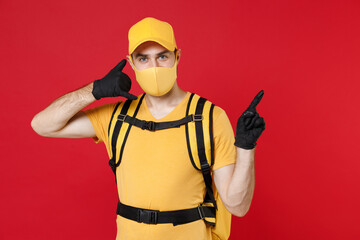Delivery employee man in yellow cap face mask gloves t-shirt uniform thermal bag backpack with food work courier service during quarantine coronavirus covid-19 virus isolated on red background studio.