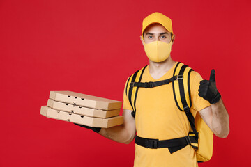 Delivery employee man in yellow cap face mask gloves t-shirt thermal bag backpack with food hold pizza in cardboard flatbox work courier service quarantine covid-19 virus isolated on red background.