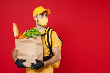 Delivery employee man in yellow cap face mask gloves t-shirt uniform thermal food bag backpack, hold paper takeaway bag products work courier service during covid-19 virus isolated on red background.