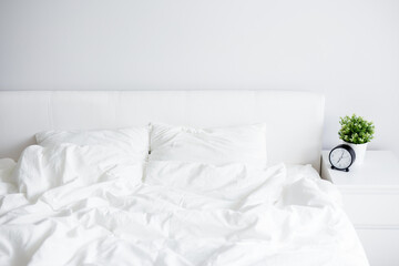 unmade bed with white blanket and two pillows, bedside table with alarm clock and home plant pot at home or hotel