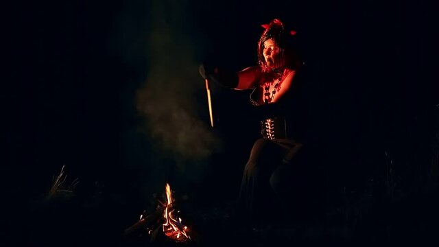 A woman in the form of a devil with horns at night by the fire with a knife dances a ritual dance in the smoke.