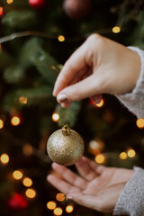 Modern glitter ornament in hands on background of festive tree in lights. Happy holidays. Woman decorating christmas tree with shiny golden bauble closeup. Preparation for christmas time.