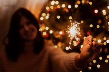 Happy woman holding firework at christmas tree with golden lights. Stylish girl with burning sparkler celebrating in festive dark room. Happy New Year