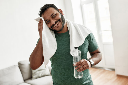 Tired but happy. Young man smiling at camera, holding water bottle and wiping sweat with towel while resting after training workout at home. Sport, healthy lifestyle