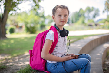 A cute happy girl with a pink backpack sits in a schoolyard or street with headphones around her neck. The children returned to school after the holidays. Shifts between lessons