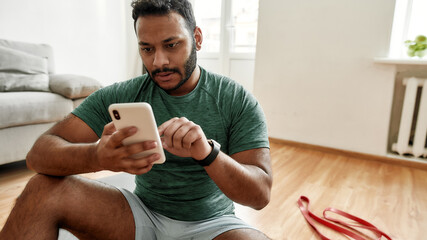 Check results. Young man using smartphone app while having morning workout at home. Freshman relaxing after training. Online personal trainer on mobile phone