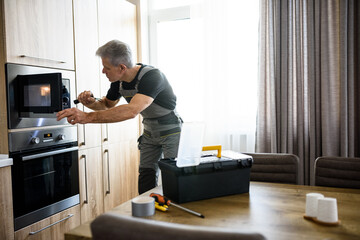Aged repairman in uniform working, examining broken microwave in the kitchen using flashlight. Repair service concept