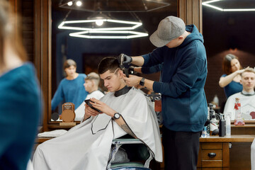 Side view of a professional barber working with hair clipper, making trendy haircut for client sitting in barber shop chair and using his smartphone