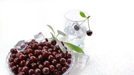 Two ripe cherries on twig in a glass with a drink, plate filled with berries in water droplets and ice cubes. Still life with tasty and healthy food close-up with copy space