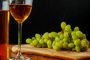 Still life of cluster of white grapes on a wooden board. Glass and bottle of white wine. Black background. Wine making concept.