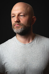Obraz premium Studio portrait of a bald man with beard in his 40s. Model dressed in simple grey t shirt looking away from the camera. Black background.