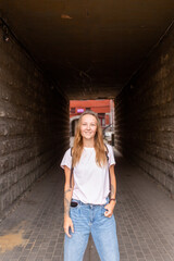 Fototapeta premium Young beautiful stylish woman sitting on the street on a Sunny summer day. A fashionable girl wears a white tshirt and blue jeans. A woman with long straight hair and tattoo
