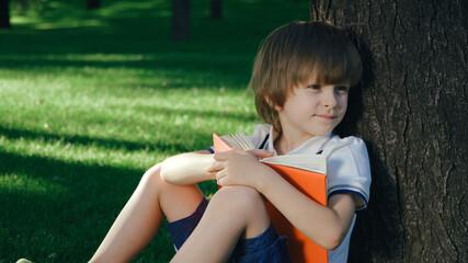 A boy is sitting under a big tree on the green grass and reading a book.