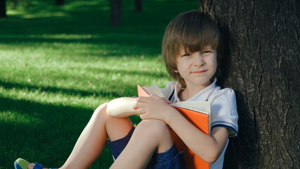 A boy is sitting under a big tree on the green grass and reading a book.