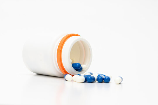 Frasco de comprimidos ca&iacute;do e aberto, saindo do seu interior capsulas nas cores azul e branca.