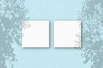 2 square sheets of white textured paper on the blue wall background. Mockup overlay with the plant shadows. Natural light casts shadows from an exotic plant.. Flat lay, top view
