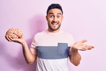 Young handsome man with beard asking for care memory holding brain over pink background celebrating...