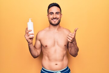 Fototapeta premium Handsome man with beard on vacation wearing swimwear holding bottle of sunscreen lotion smiling happy and positive, thumb up doing excellent and approval sign