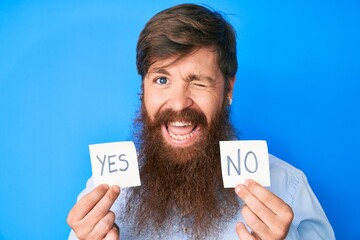Handsome young red head man with long beard holding yes and no reminder winking looking at the...