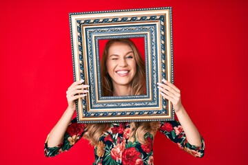 Young caucasian woman with blond hair holding empty frame winking looking at the camera with sexy...
