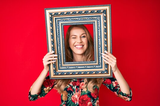 Young caucasian woman with blond hair holding empty frame winking looking at the camera with sexy expression, cheerful and happy face.