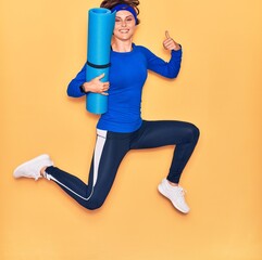Young beautiful sportswoman smiling happy. Jumping with smile on face holding yoga mat doing ok sign with thumbs up over isolated yellow background.