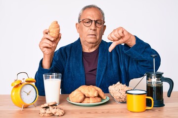 Senior handsome man with gray hair sitting on the table eating croissant for breakfast with angry face, negative sign showing dislike with thumbs down, rejection concept