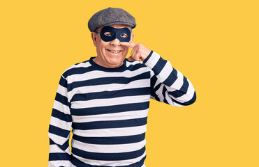 Senior handsome man wearing burglar mask and t-shirt pointing with hand finger to face and nose, smiling cheerful. beauty concept