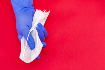 Medical mask, covid-19, a hand in a blue medical disposable glove clutches a white mask on a red background, the concept of the end of the coronavirus.