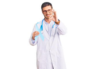 Fototapeta premium Handsome young man with bear wearing doctor uniform and holding blue ribbon doing ok sign with fingers, smiling friendly gesturing excellent symbol